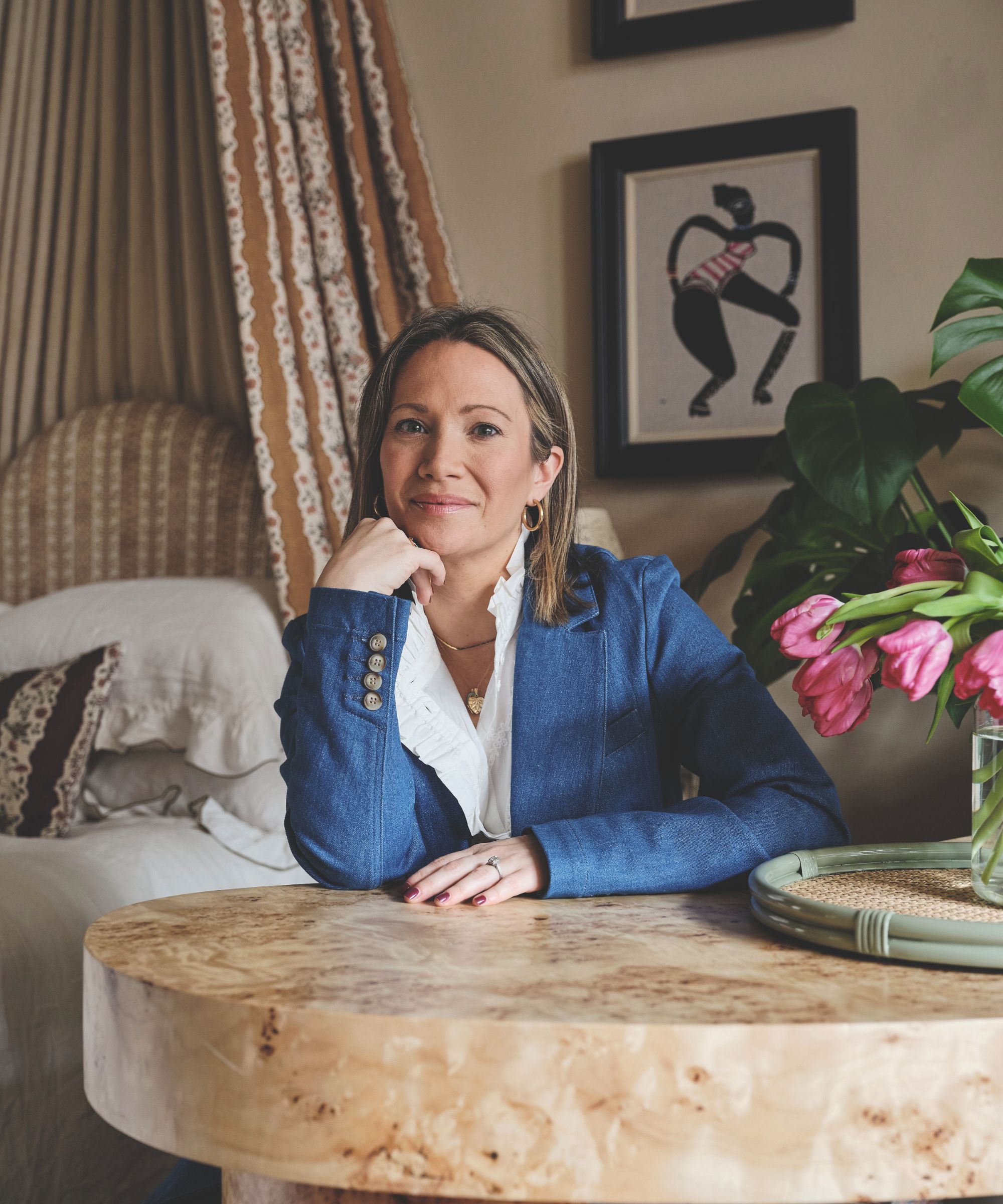 A woman in a blue blazer sits at a table in a bedroom. 