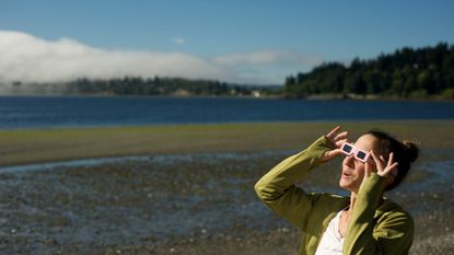 A woman wears eclipse glasses while standing on a beach