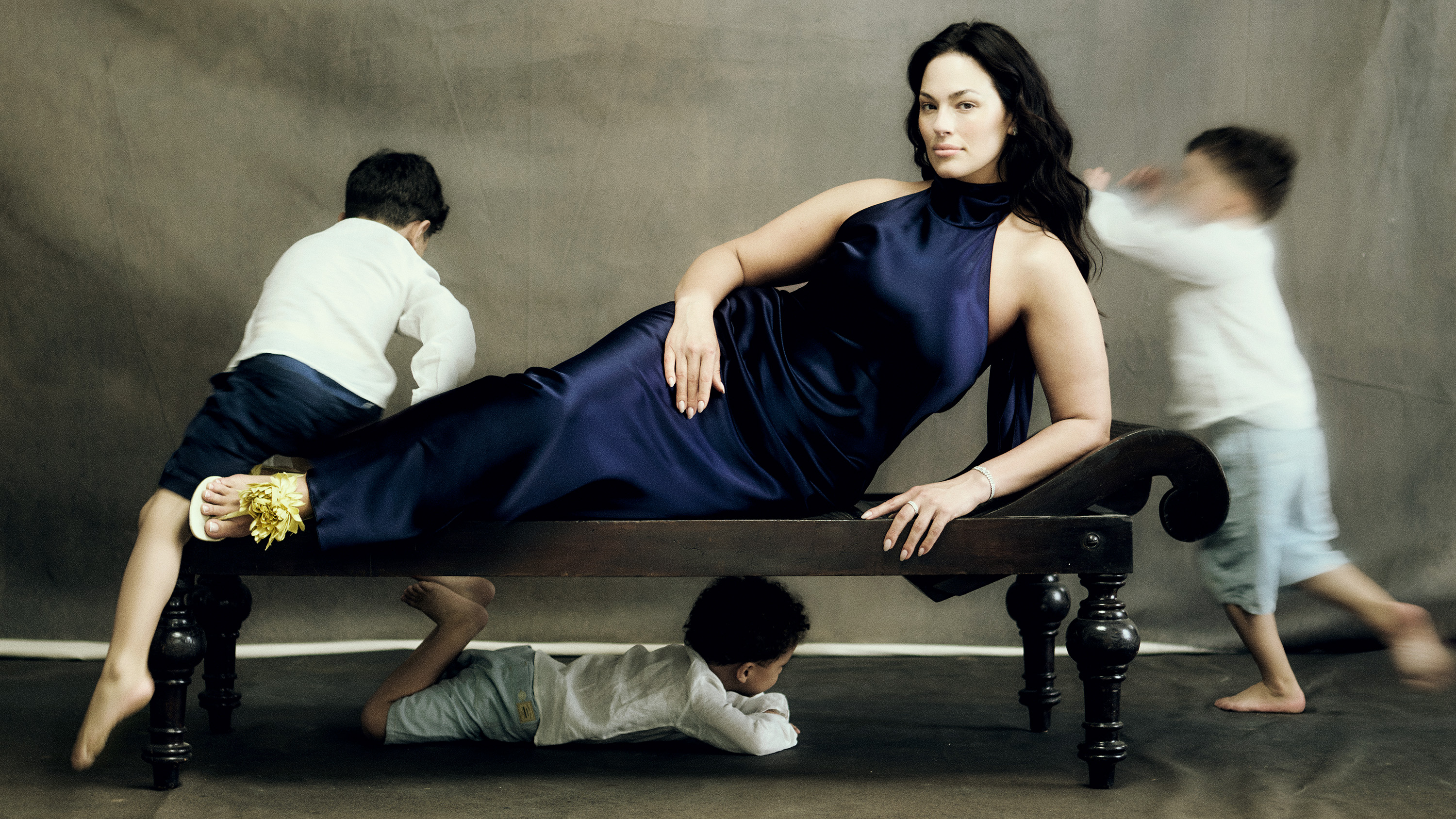 Ashley Graham wears a blue satin dress lying on a couch as her three sons run around her 