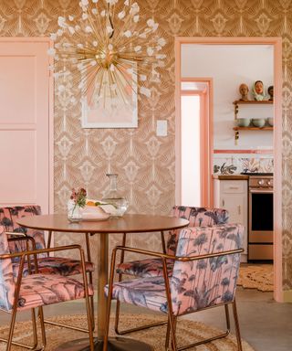 A dining room with patterned wallpaper, a gold chandelier, pink doors, and a wooden circular dining table with four palm leaf patterned pink dining room chairs around it