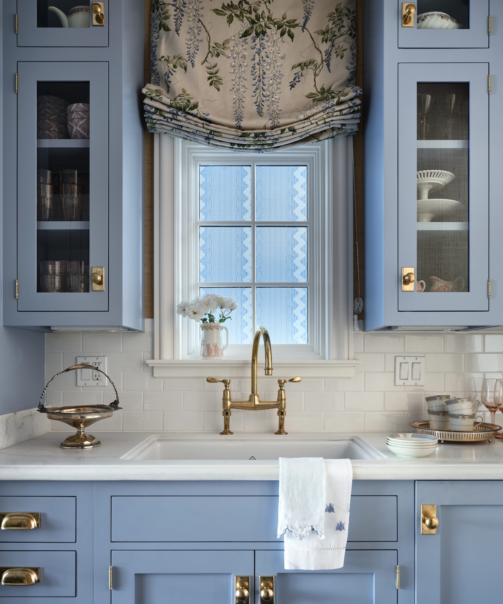 Blue pantry cabinetry with glass-fronted cupboards, a marble sink, brass tap and a floral Roman shade.