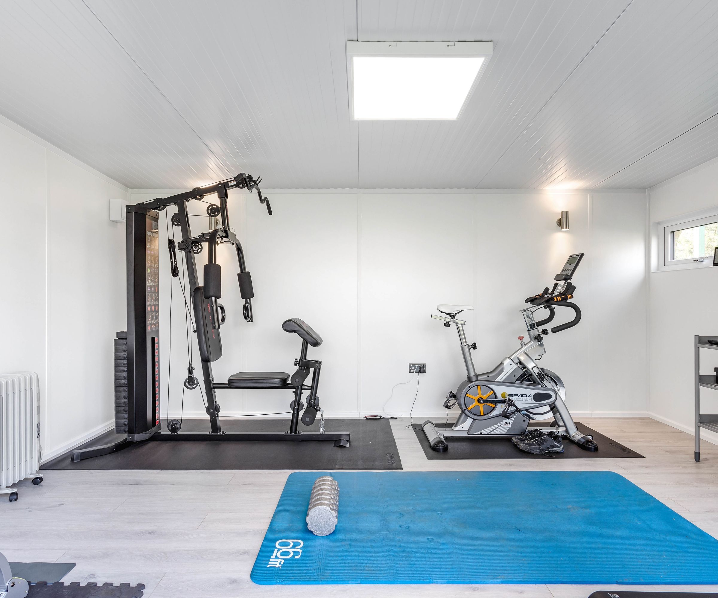 Home gym with weight machine, spin bike, blue exercise mat and dumbbells on light wood flooring.