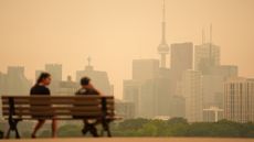 Two people sit on a wooden bench in front of a brown, smoggy cityscape