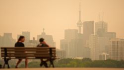 Two people sit on a wooden bench in front of a brown, smoggy cityscape