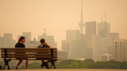 Two people sit on a wooden bench in front of a brown, smoggy cityscape