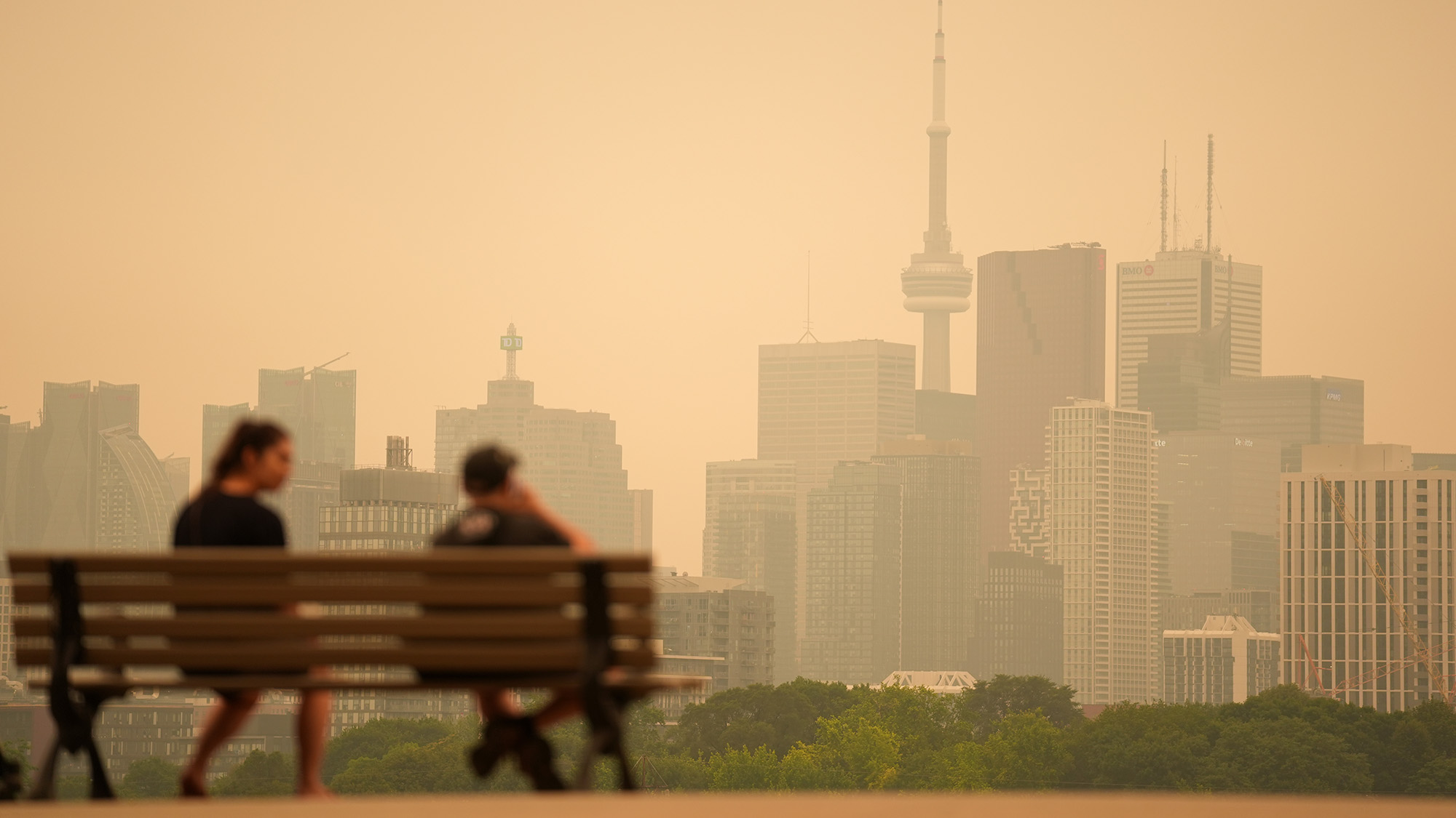 Two people sit on a wooden bench in front of a brown, smoggy cityscape