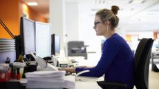 white woman blonde hair bun tied blue top sitting in office on chair hands on keyboard looking at computer screen papers on desk sideways to camera