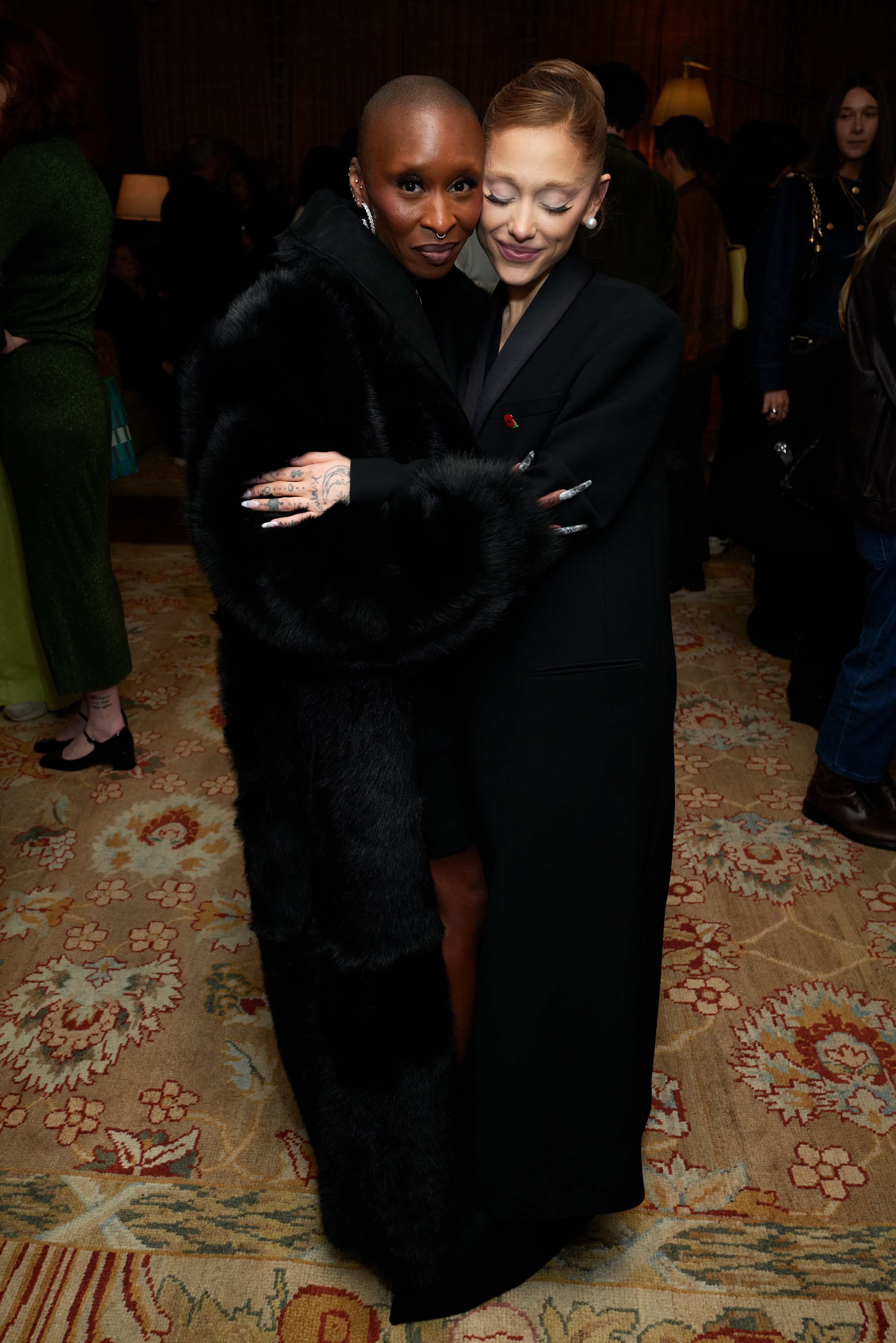 Ariana Grande hugging Cynthia Erivo as they pose for a picture. Both are wearing long black coats.