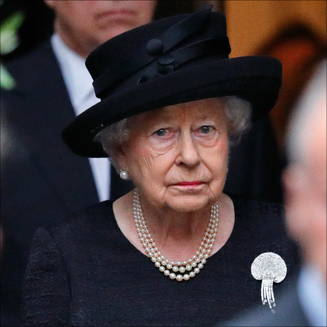 Queen Elizabeth II wears a black dress and matching hat and looks very sad while wearing a string of pearls