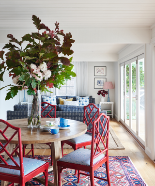 dining and living area with red vintage style chairs and round modern wooden table and patterned rug and blue patterned sofa in back drop and big vase of flowers on table