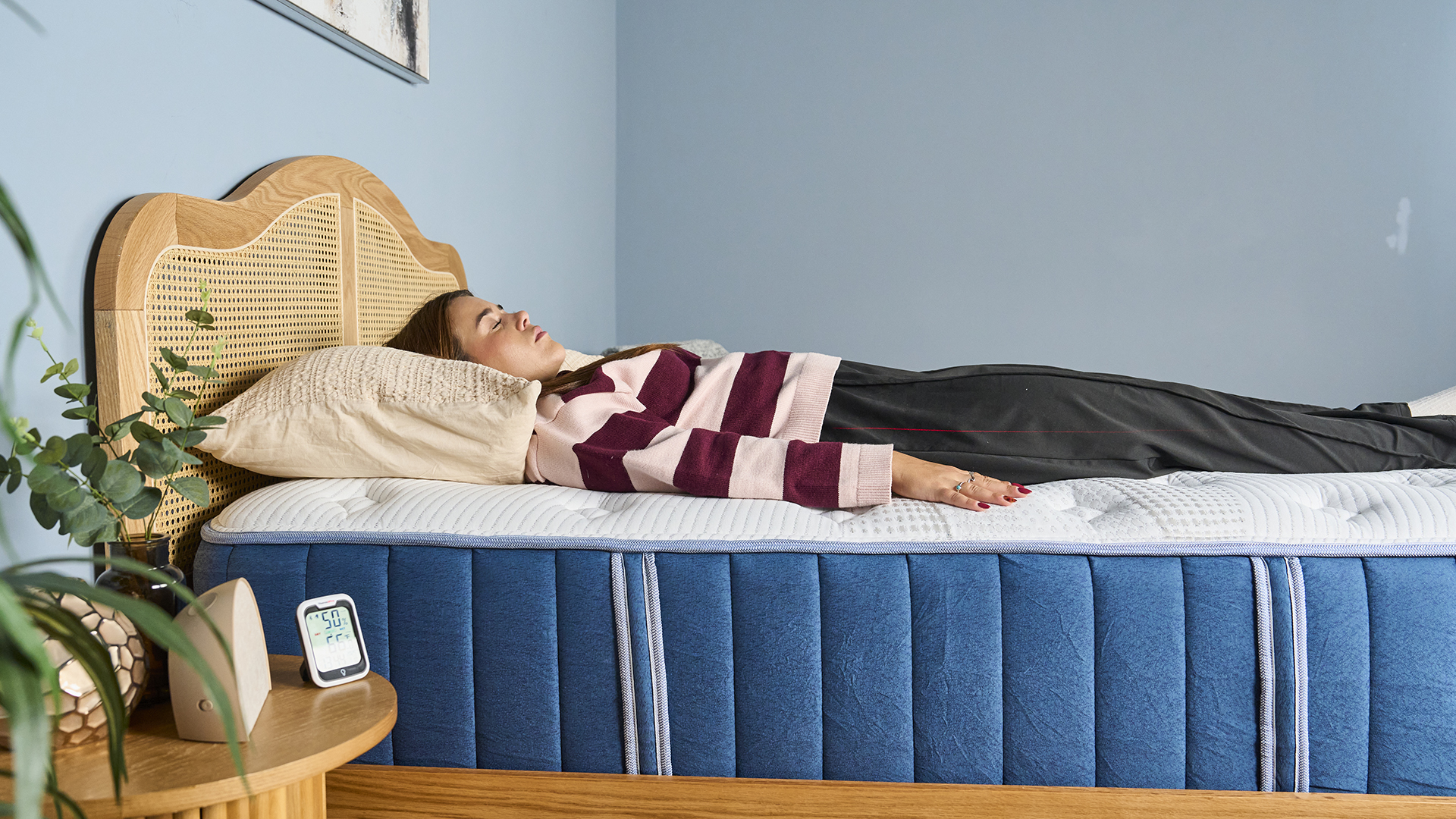 Side view of our mattress tester wearing a striped jumper and black trousers laying on her back on the navy and white Bear Elite Hybrid mattress on a wooden bedframe with a rattan headboard. Next to a bedside table with a sunrise alarm clock and room temperature thermometer and green plant.