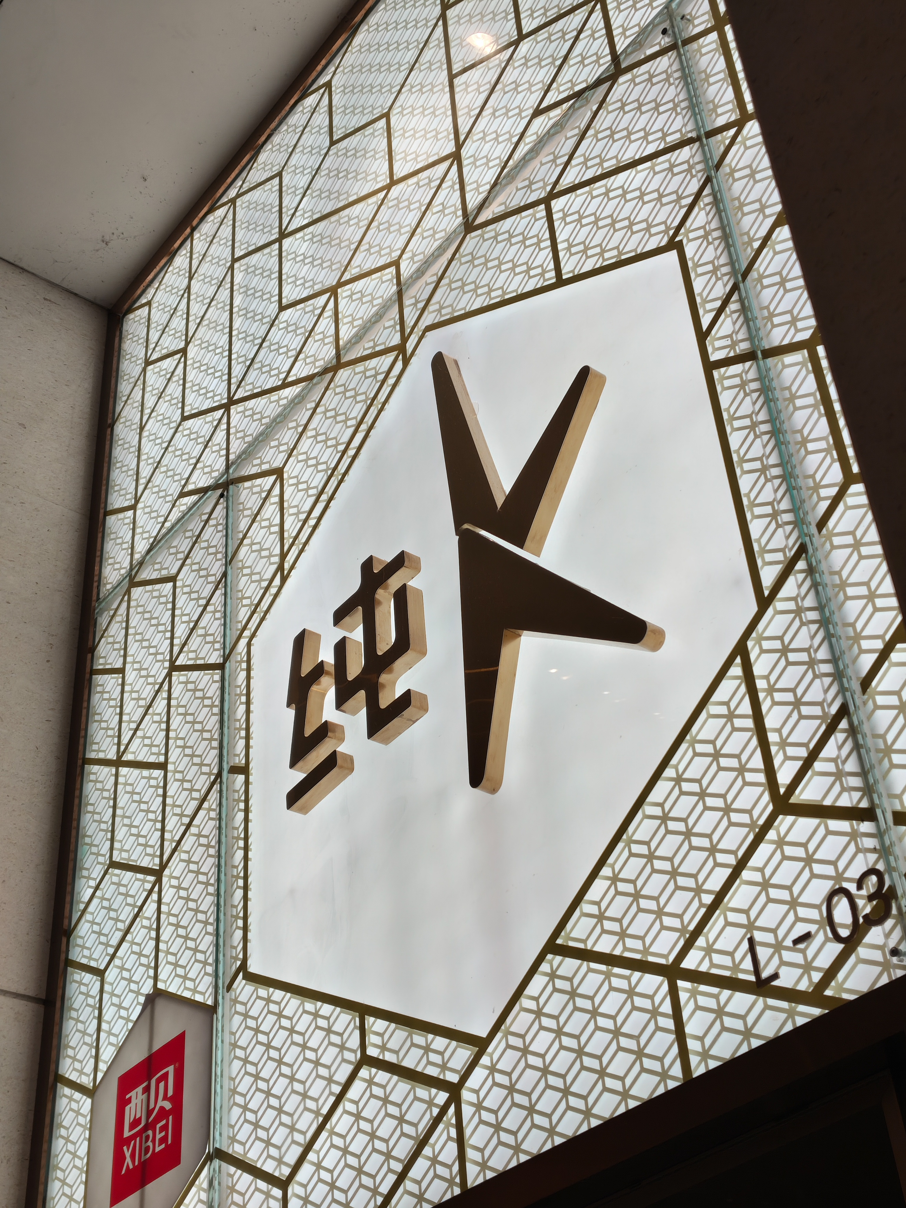 A close-up shot of a modern illuminated sign. The sign features a stylized, dark gold letter "K" and Chinese characters on a white hexagonal background. The background is surrounded by a frosted glass panel with an intricate gold geometric pattern.