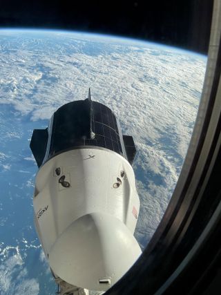 through a rounded window looking downward, a white space capsule is seen with dark solar panels lining its aft trunk. Below, the blue of Earth, with a wash of white clouds.
