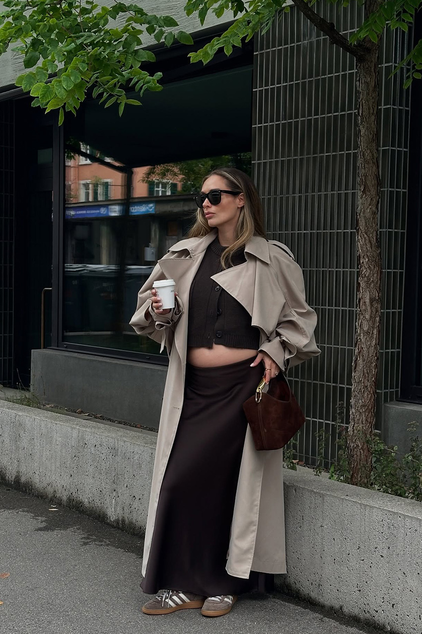 How to wear a slip skirt is shown in a photo of a woman&#039;s slip skirt outfit with a woman standing outside wearing a sunglasses with a long trench coat over a brown cardigan with a matching slip maxi skirt, suede samba sneakers, and a brown suede handbag