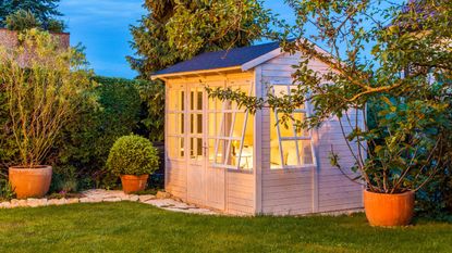 A white garden shed with large, open windows. Painted white. In a back yard fenced in with tall bushes, potted trees, and shrubs around the shed. A small stone garden path leading to the shed through short grass lawn