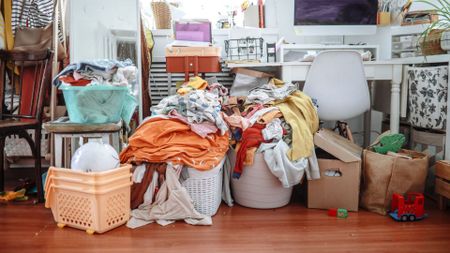 A cluttered bedroom with wood floors showing laundry baskets piled up with clothes and random toys and objects on the desk and floor