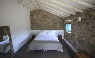 Bedroom with bed with white linen on wooden floor. There is one white wall and a cement and brick wall with a window