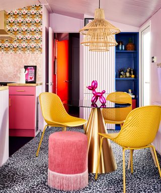 A pink living area with a brushed gold table with two yellow seats either side, a rattan pendant light above it, and a blue leopard print carpet underneath it