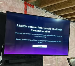 A TV mounted on a wall with the Netflix password crackdown message: "A Netflix account is for people who live in the same location Everyone who lives at your primary location can use Netflix when they're on the go or traveling. If this is your account, we'll help you set your primary location on the next screen." and a Continue button.