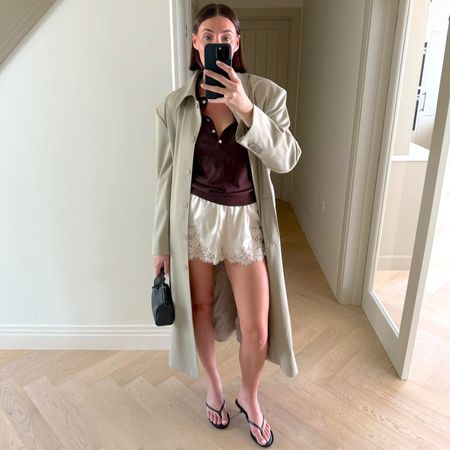 British style influencer Sarah Louise Blythe poses for a mirror selfie wearing a trench coat, brown top, satin lace-trimmed shorts, mini black bag, and black flip-flop kitten heel sandals