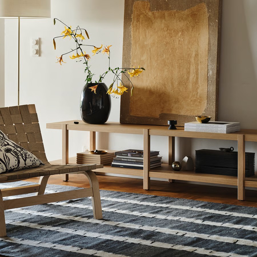 Image of the corner of a white living room with hardwood floors and a navy blue and white striped rug. There is a long and low storage unit against the wall with a gold abstract art piece, a black vase, and stacks of books on it. There is a beige, woven chair in the room as well.