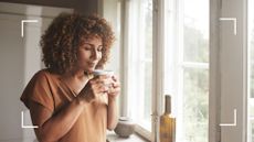 Woman standing smiling with cup of tea or coffee by window at home, representing how to stop drinking alcohol for good 