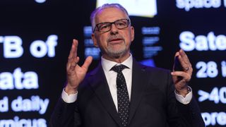 a man in a black suit and glasses holds his hands up at shoulder height while speaking on a stage