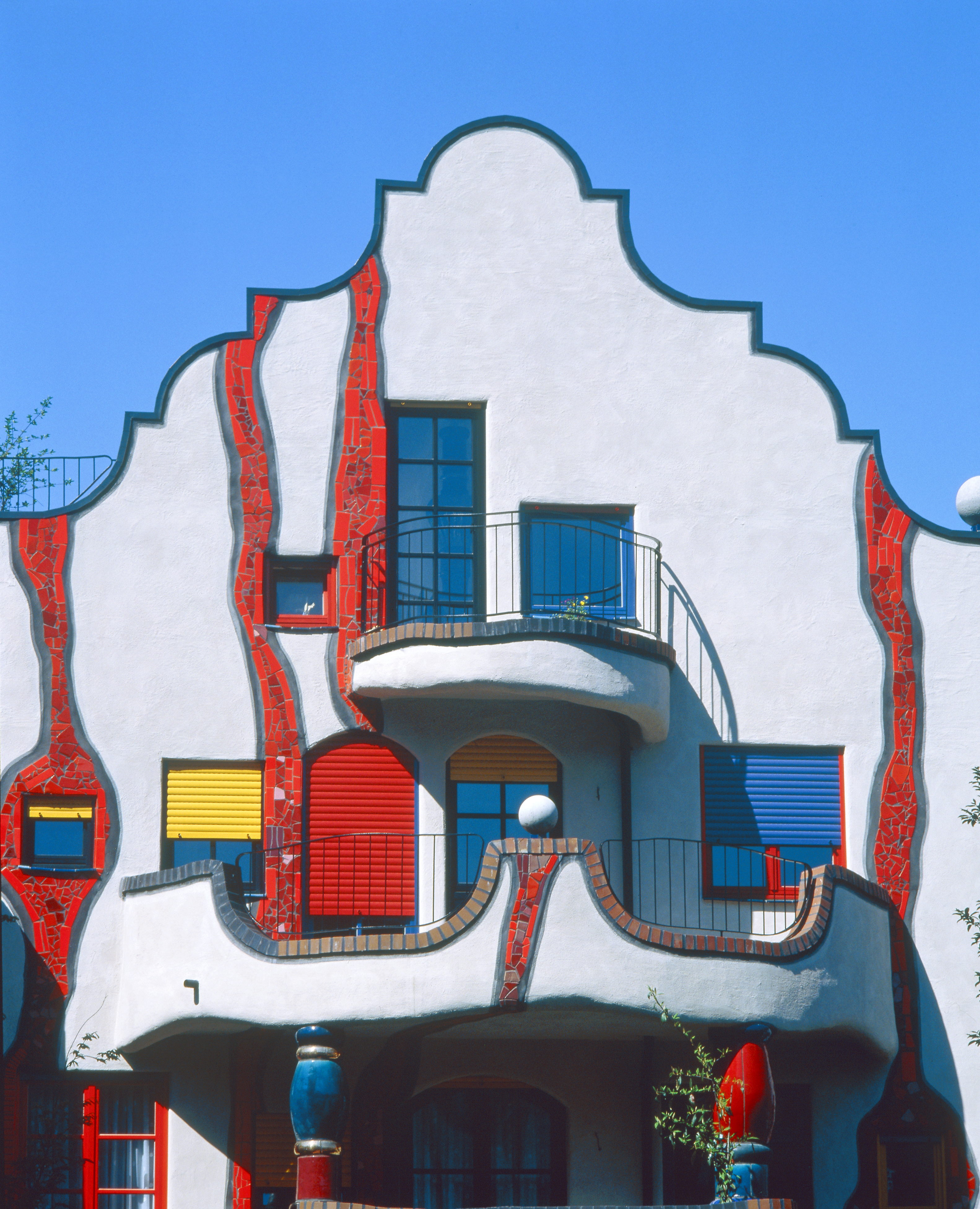 The colorful, wiggly facade of a building with a white surface brought to life by red glazed tiling, yellow, blue, and red window blinds, and wrought iron balconies.