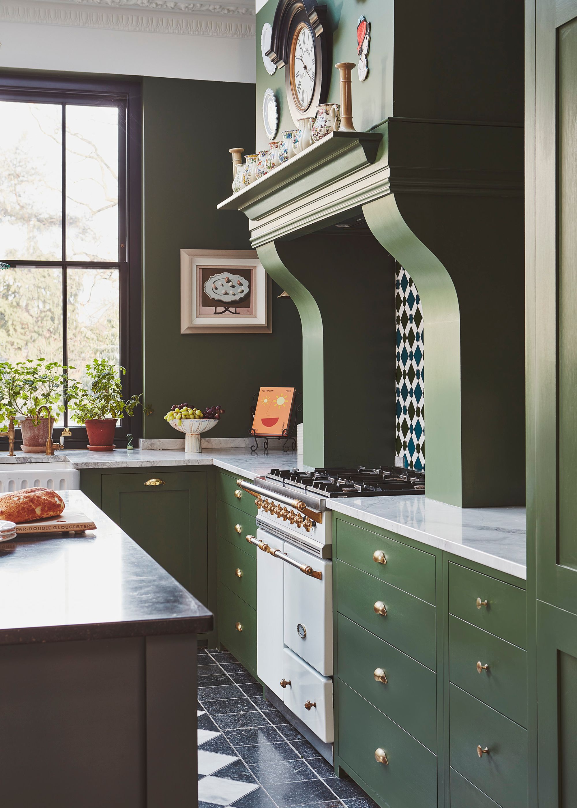A deep green kitchen with a marble white countertop and a dark island opposite it. On top of the worktops are a bunch of accessories as well as plants in plant pots