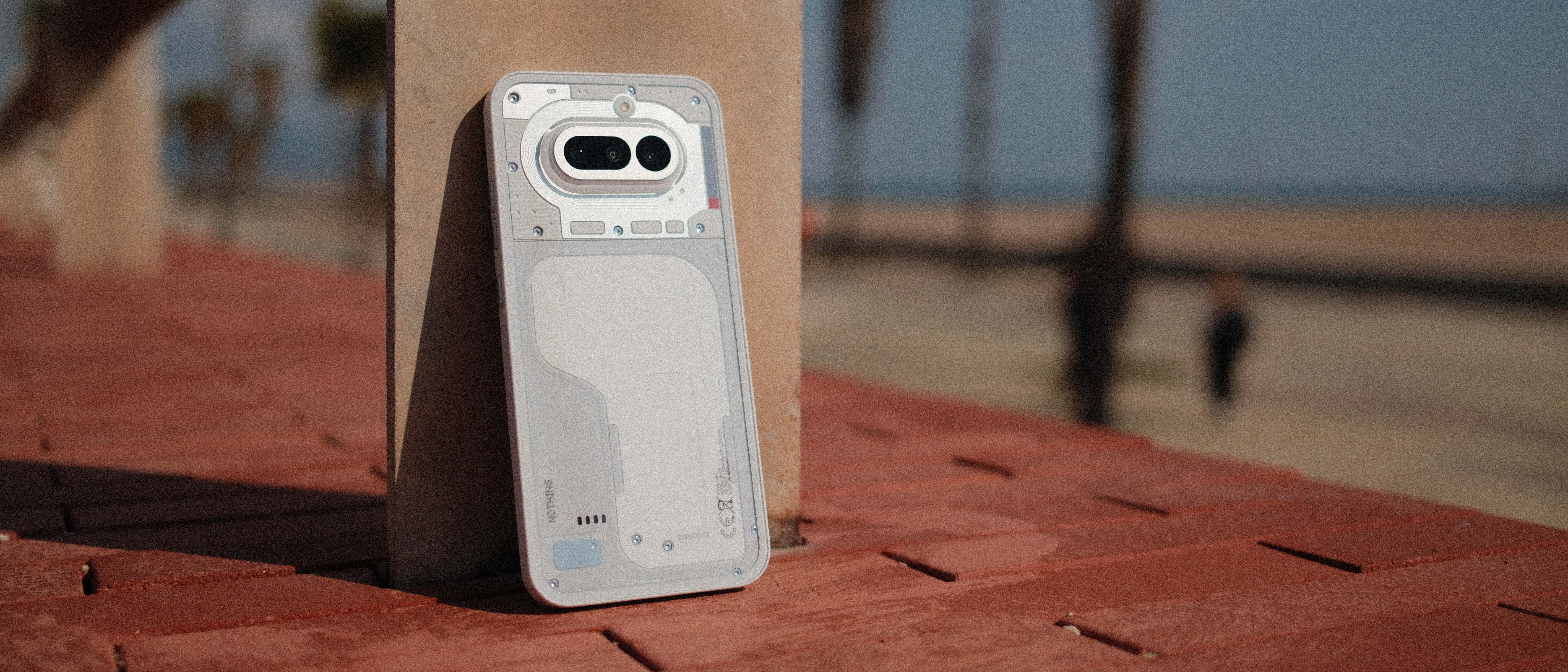 Nothing Phone (4a) in white standing against a pillar on a brick surface at a beach promenade, showing its transparent-style rear design and dual camera module.