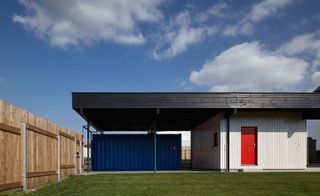 The roof residential home clad in black timber planks with the main body of the house in white wood with a red door. A blue container is one the left of the house shielded by the timber plank roof