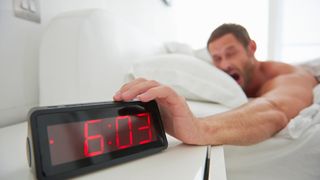 Man yawning and snoozing an alarm clock that reads 6am