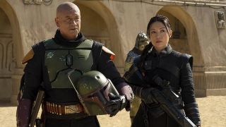 A bald man in armor carrying his helmet, walking beside a woman in black carrying a gun.