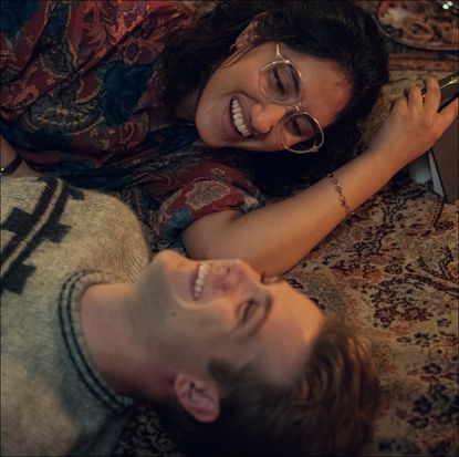 a woman (Ambika Mod as Emma) holds a book as she and a man (Leo Woodall as Dexter) lie on the floor while laughing