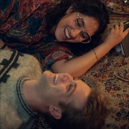 a woman (Ambika Mod as Emma) holds a book as she and a man (Leo Woodall as Dexter) lie on the floor while laughing