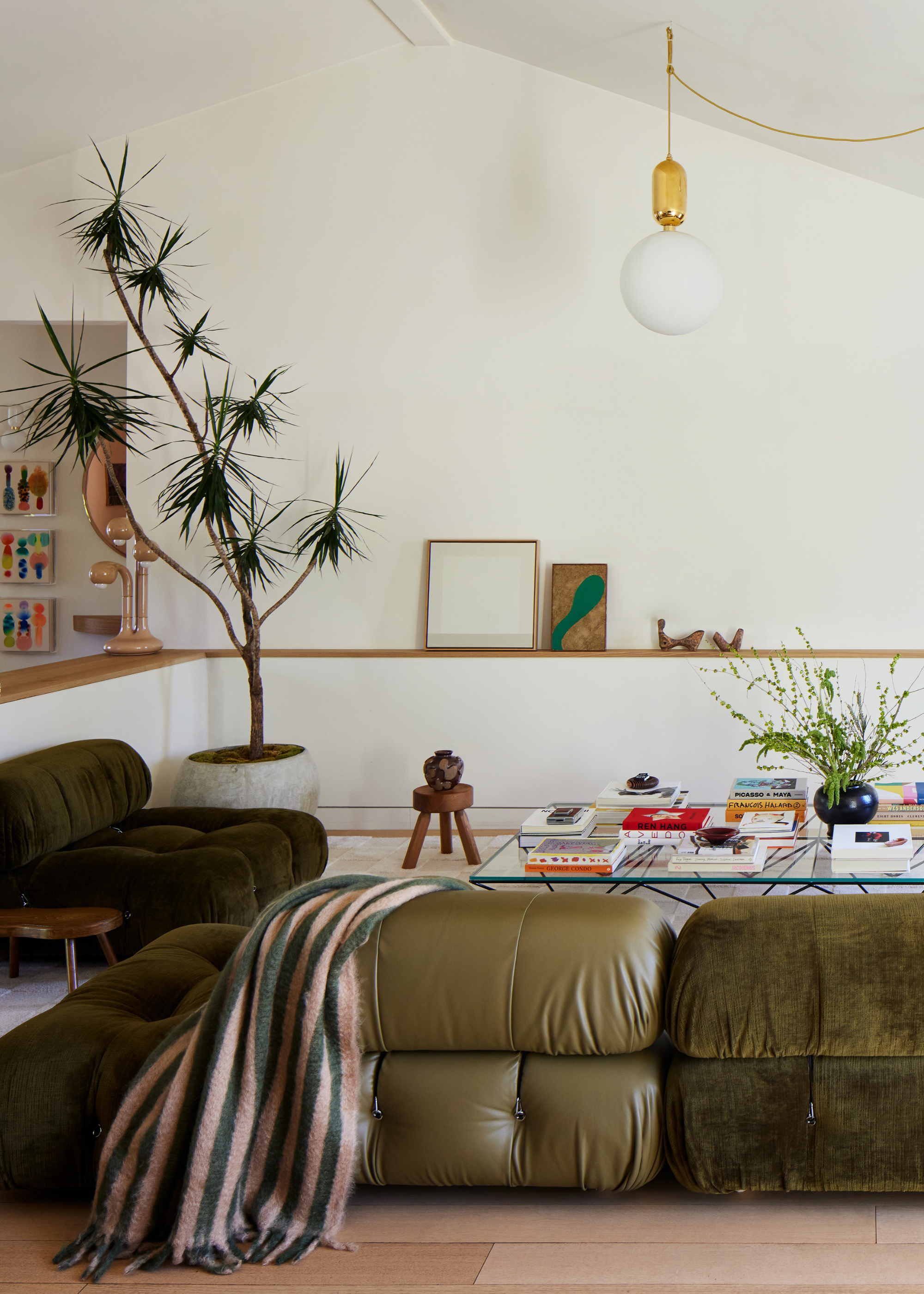 A white living room with a potted tree, a green leather and suede sofa set, a wood stool with a vase, a transparent coffee table with books and a plant, alongside a shelf with standing art pieces