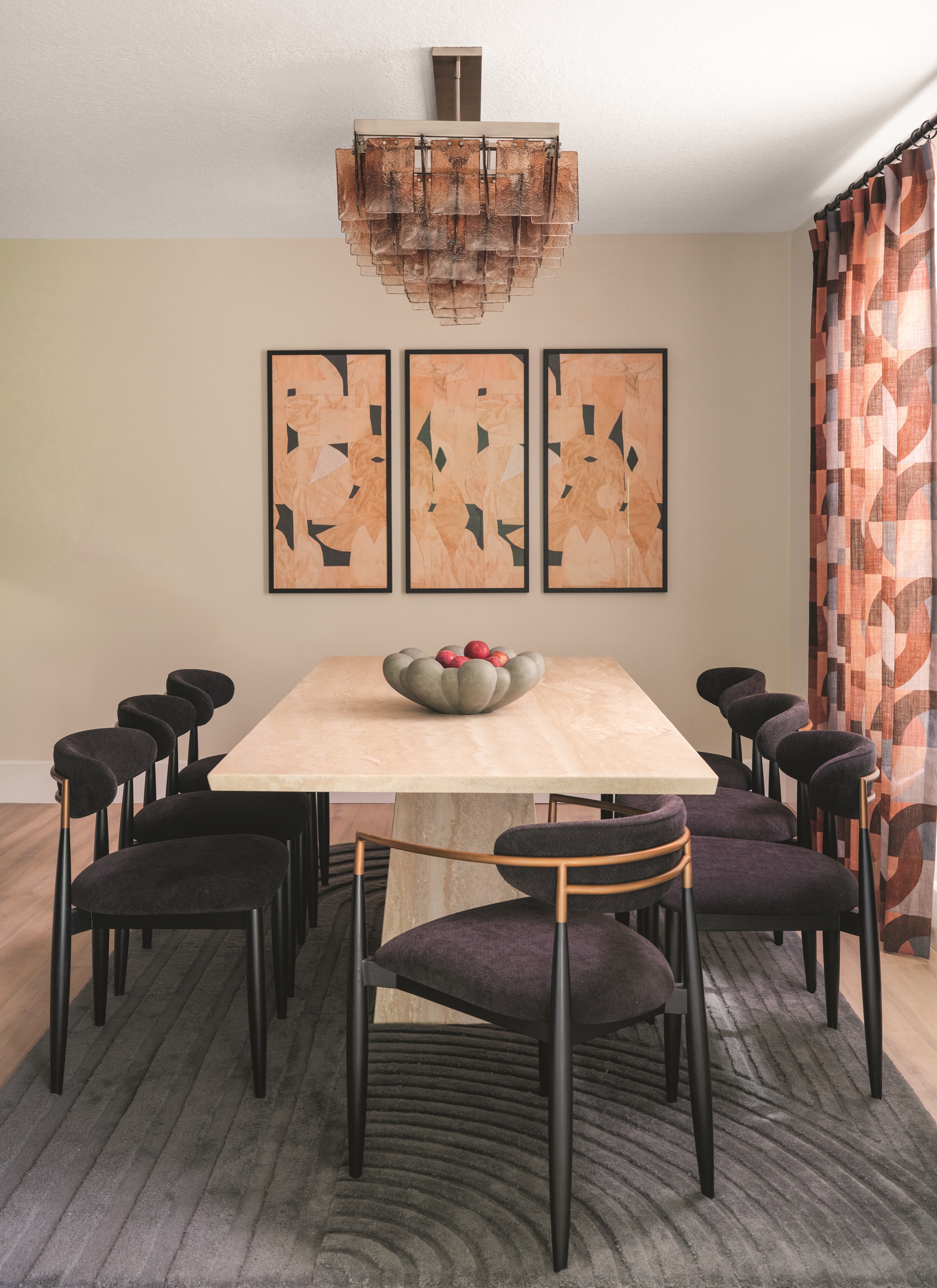 a dining room with a travertine table and black dining chairs with patterned curtains and a trio of wall art