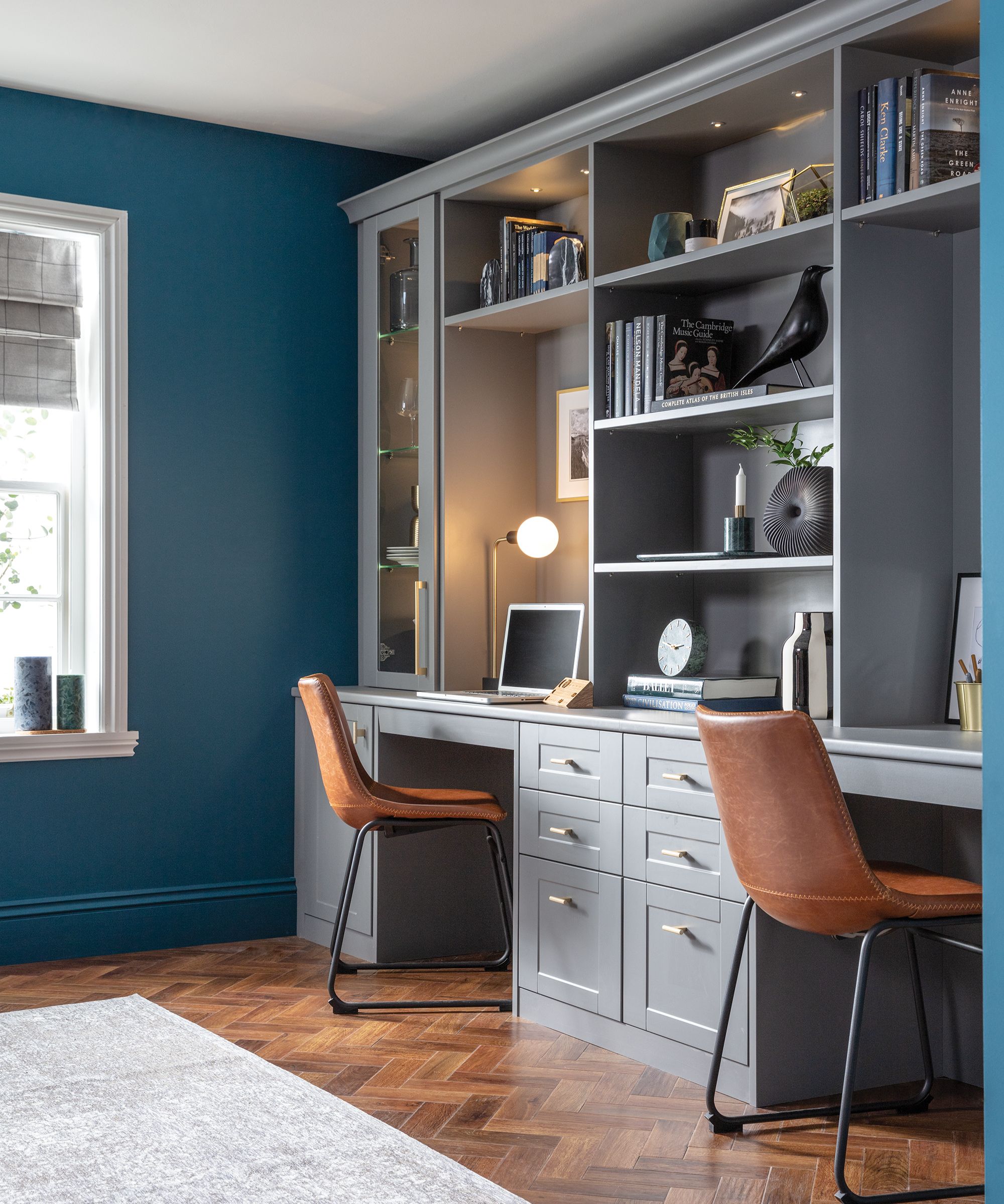 A smart built-in double desk with grey cabinetry, leather chairs, and styled shelving sits against rich teal walls and parquet flooring.