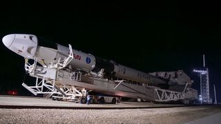 a white rocket and space capsule are transported to the launch pad horizontally at night.