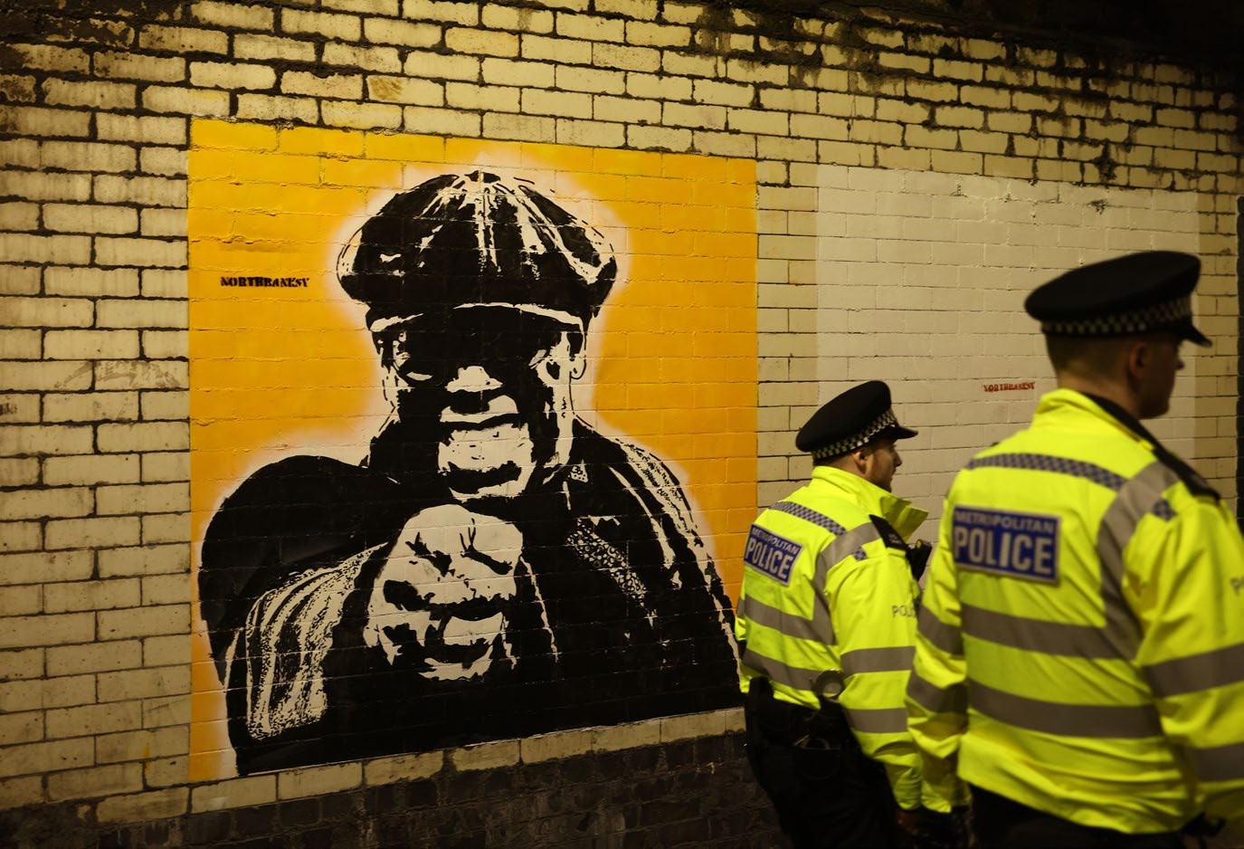 A striking mural of a man pointing, set against a vibrant yellow background, alongside two police officers in reflective jackets in a tunnel, reflecting the intersection of street art and community.