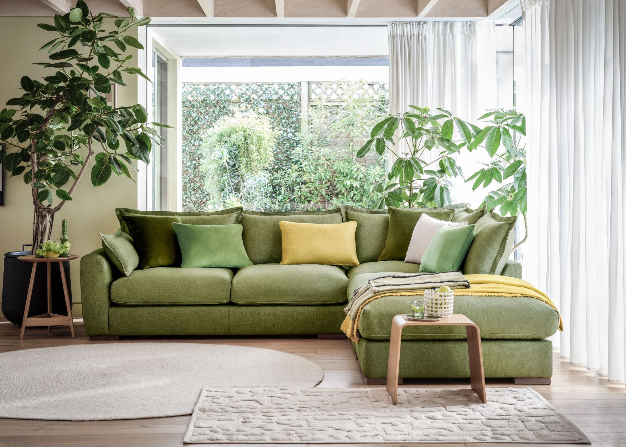 modern living room with green corner sofa, yellow and green accent cushions, houseplants and sliding glass doors with sheer curtains