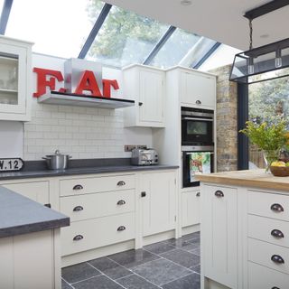 An L-shaped kitchen with cream cupbaords and black top, large angled skylights, exposed brick wall in the corner and a red E A T sign on top of the extractor fan