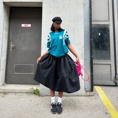 Woman wears black Adidas Samba sneakers, full black skirt, Adidas soccer jersey and a page-boy cap.