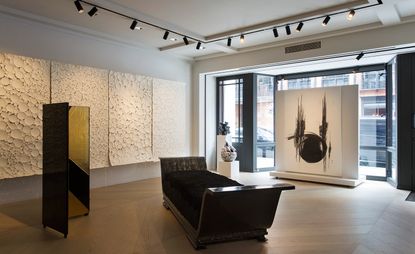 Katsu Hamanaka's work is displayed at the Dutko Gallery. In the middle of the room, there is a dark wood and black fabric sofa, with a privacy screen in dark brown with golden details to the left. An abstract painting in black & white is hung to the right.