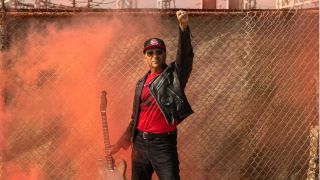 Tom Morello raiding his fist, holding a guitar, in front of a chain link fence as red smoke swirls around him