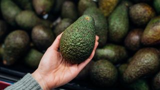 Woman's hand holding an avocado in front of a load of avocados sat together, perhaps at a supermarket