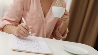 Middle aged woman with cup of drink solving sudoku puzzle at white table indoors, closeup