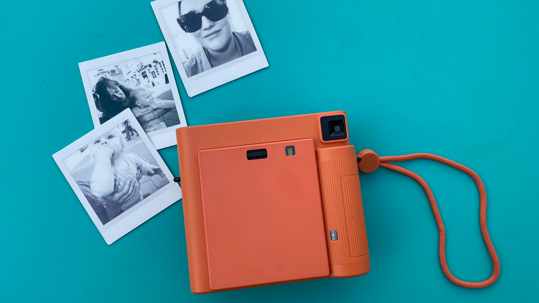 Orange instant camera with wrist strap next to three black and white instant photos on a teal background