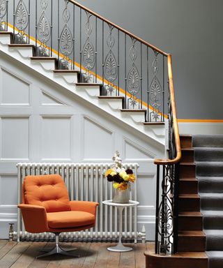 Slate painted stairwell with painted orange accent stripe, under-stair wall paneling painted white, dark wooden stairs with gray carpet runner, dark wooden flooring with bright orange lounge chair, white tulip table with vase of flowers