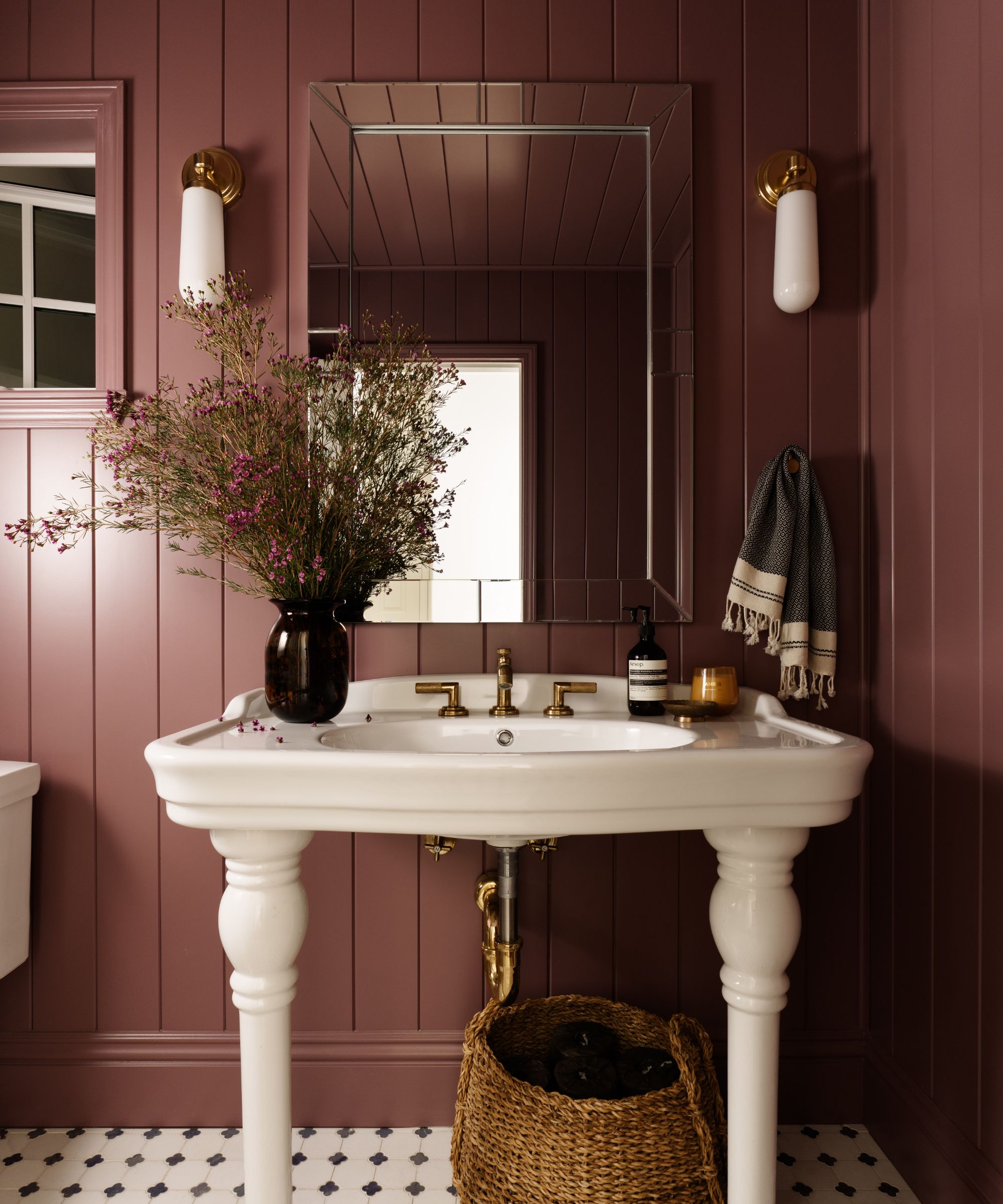 A small powder room with burgundy panelled walls, a white freestanding sink with brass hardware, a vase of flowers, a mirror with two wall sconces on either side, and black and white tiled flooring.